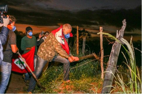 A group of people, some wearing masks, gather at dusk as one person uses a wooden stick to break a barbed wire fence. One person holds a red flag with a symbol.