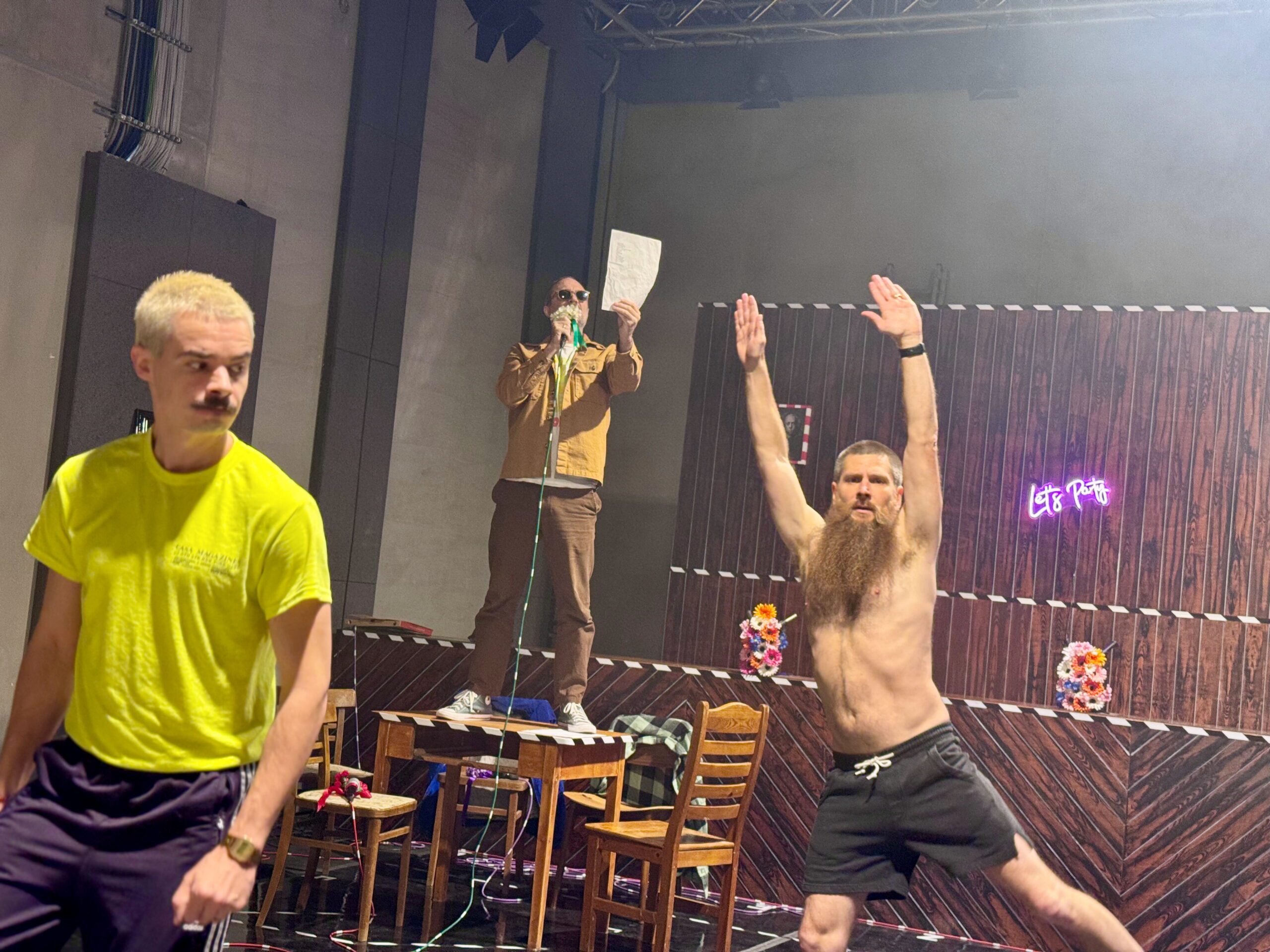 Three men perform on stage: one in a yellow shirt walks, one shirtless with a long beard raises arms, and another stands on a chair reading from a paper. The background features a neon sign and wooden panels.