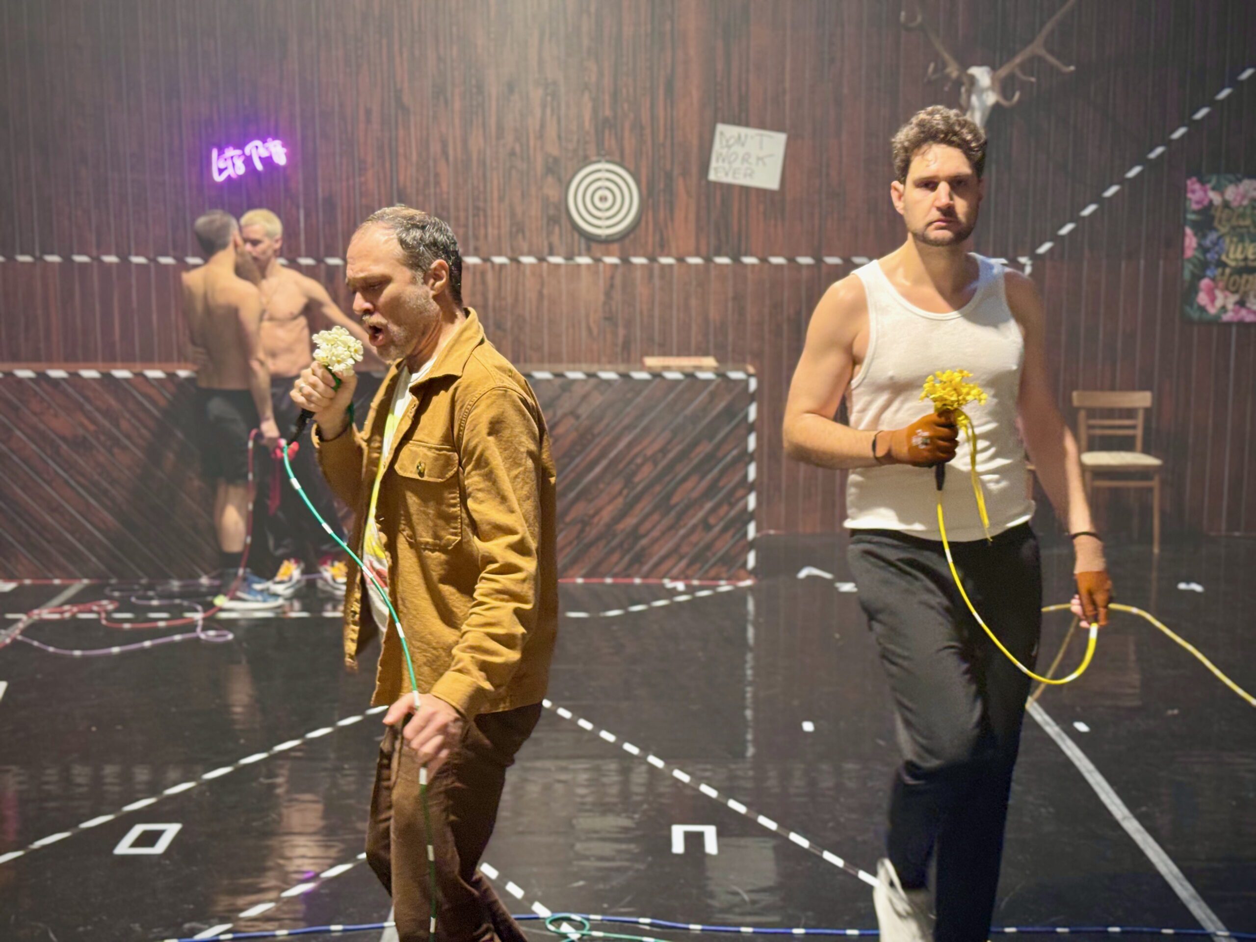 Two men holding yellow flowers and wired microphones stand in a marked area. Two shirtless men face each other in the background. Neon signs, flowers, and a mounted deer head decorate the wooden walls.