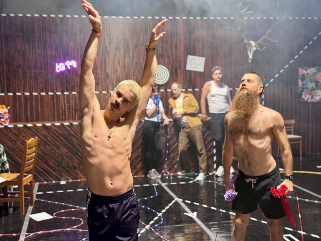 Two shirtless men pose in a room with wooden walls, scattered chairs, darts on the wall, and a mounted deer head. One man raises his arms; the other has a long beard and holds flowers. Others stand in the background.