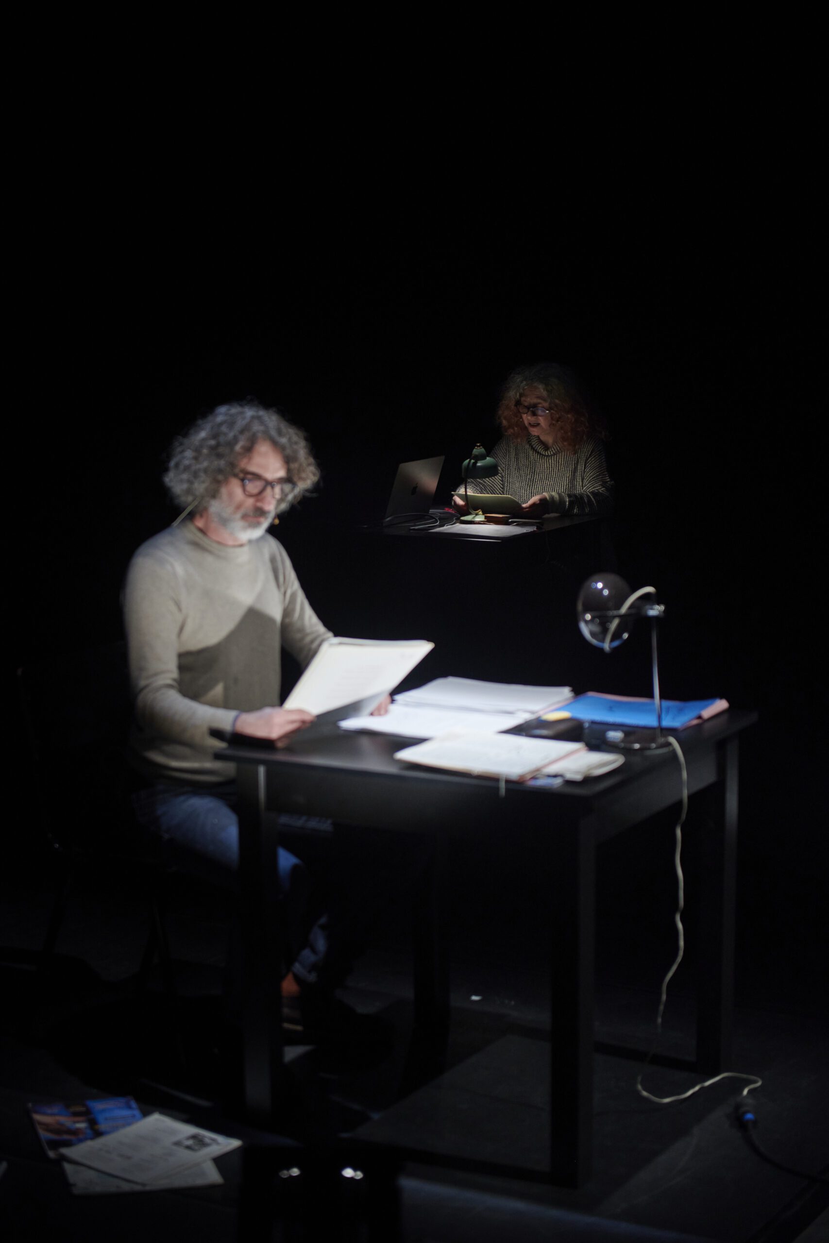A man with curly gray hair and glasses sits at a desk reading papers, illuminated by a lamp, with a smaller reflection of him working at a computer projected above and behind him in the dark.