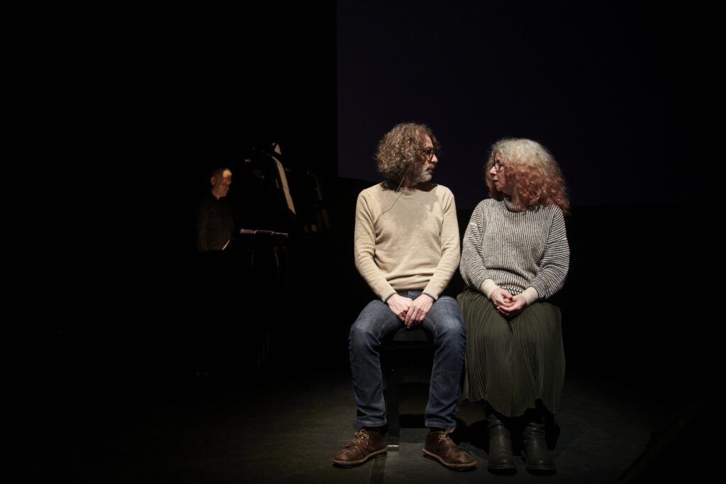 Two people with curly hair sit side by side on a dark stage, facing each other. A person at a piano is dimly visible in the background, partially out of the light.