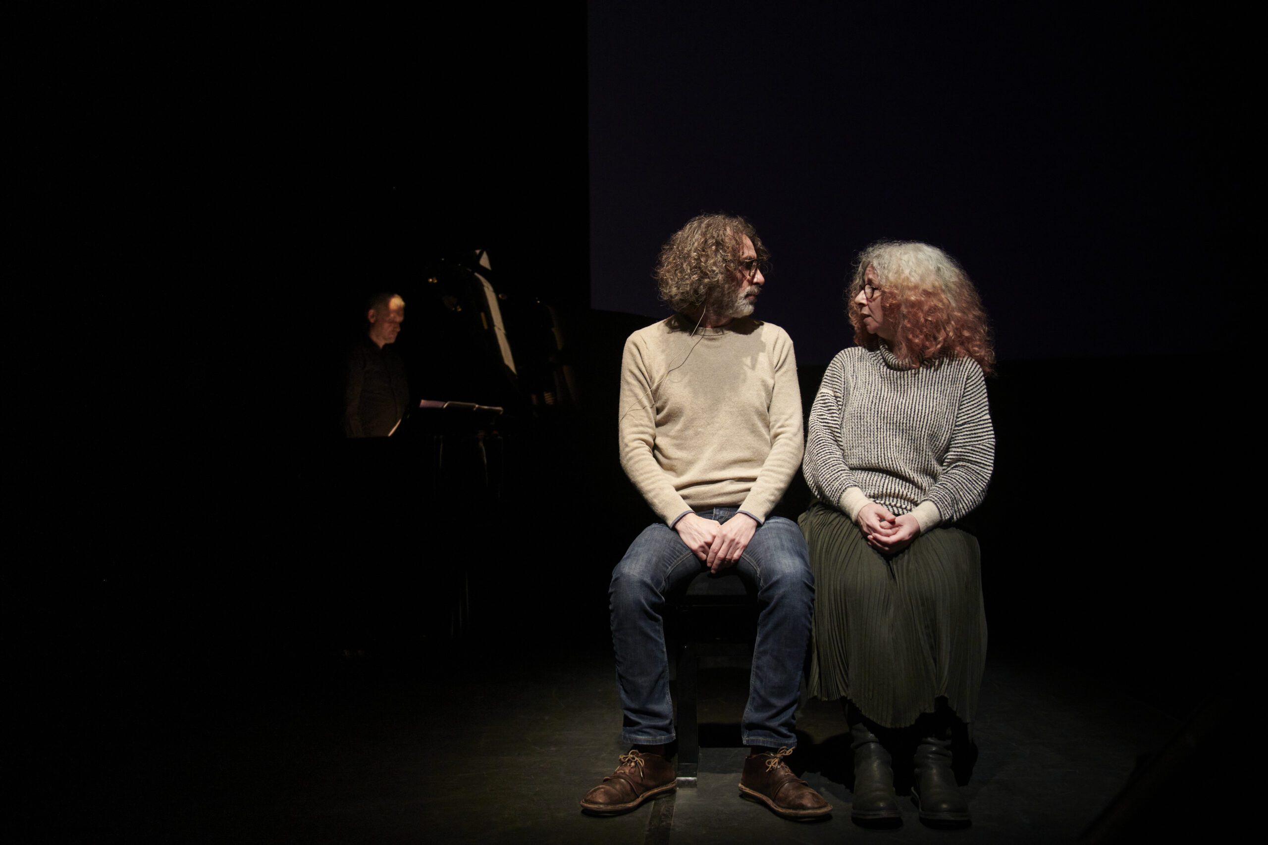 Two people with curly hair sit side by side on a dark stage, facing each other. A person at a piano is dimly visible in the background, partially out of the light.