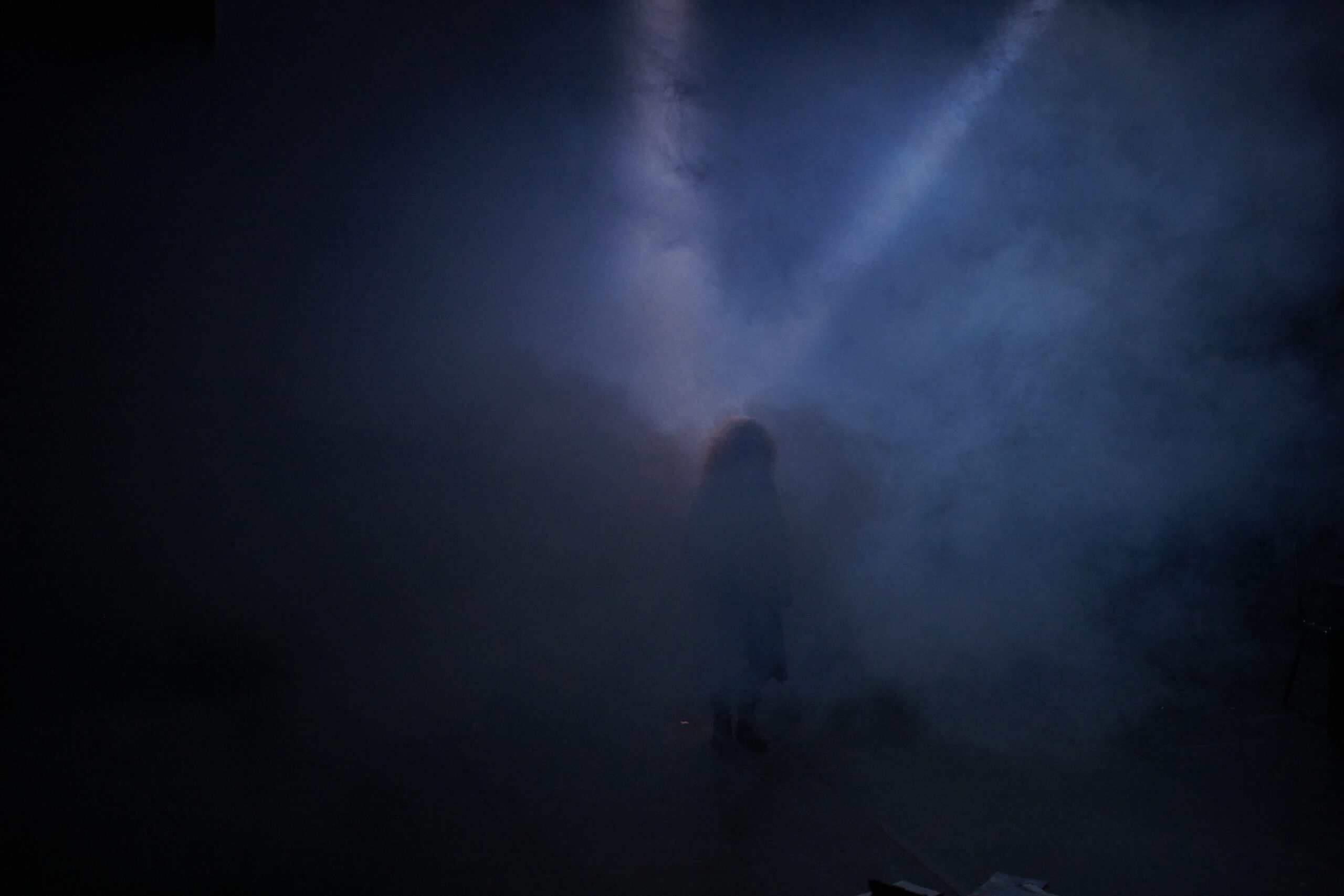 A person stands in heavy fog, partially silhouetted by a dramatic beam of blue-tinted light shining down through the mist, creating a mysterious and atmospheric scene.