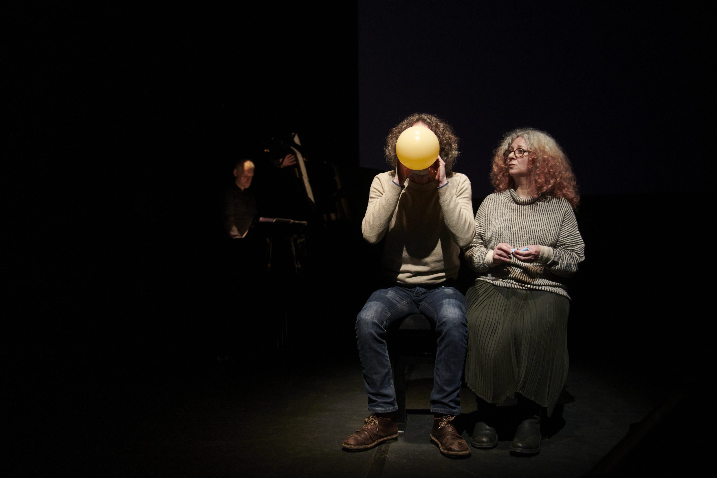 Two people sit on stage in dim lighting; one holds and looks at a yellow balloon, the other watches. A musician plays a keyboard in the background. All wear casual clothing.