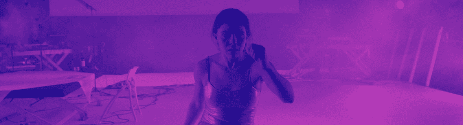 A person in a shiny outfit stands in a purple-lit, foggy room with musical equipment and chairs in the background, looking toward the camera.