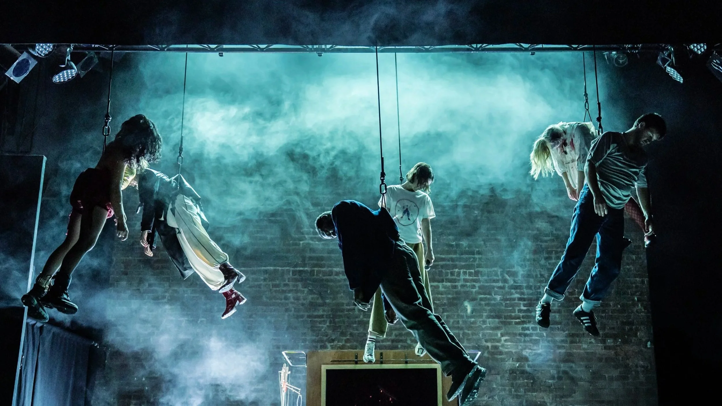 Five people are suspended by harnesses on stage in dramatic lighting and mist, appearing to hang limply in front of a brick wall, creating a haunting, theatrical scene.