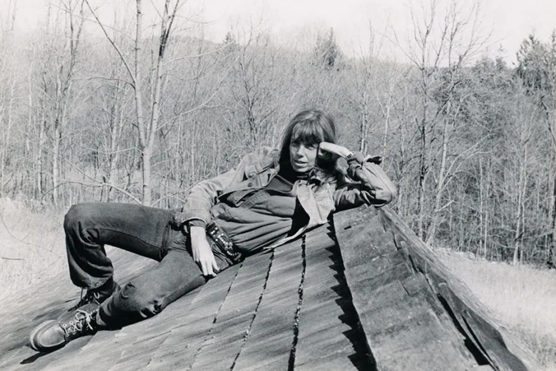 A person with long hair, jeans, and boots reclines on a slanted rooftop, resting their head on one hand. Leafless trees fill the background in a natural outdoor setting.