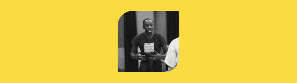 A person gestures passionately while speaking, wearing a dark T-shirt and standing indoors. The photo is black and white and placed on a solid yellow background.