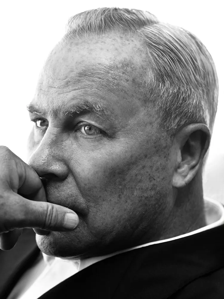 Black and white close-up of a man in a suit, gazing thoughtfully to the side, with his hand resting near his mouth.