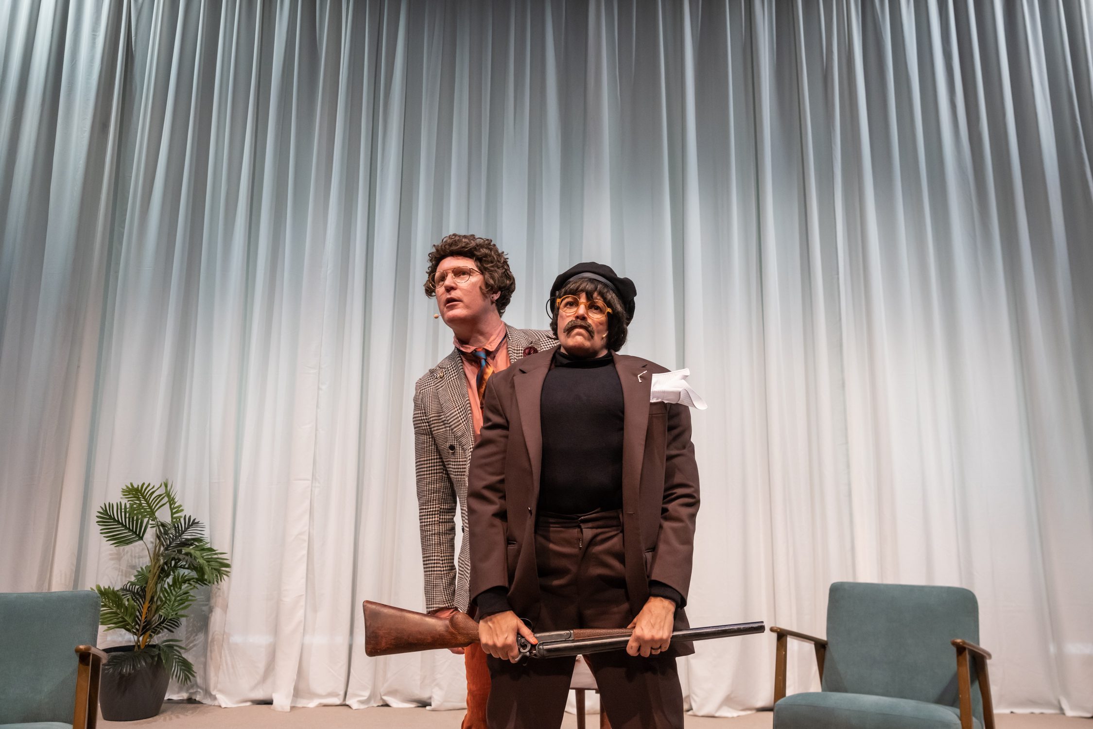 Two people in costume stand in front of white curtains. One wears a wig and plaid suit, the other has a mustache, beret, fake eyebrows, and holds a prop rifle. Two chairs and a potted plant are also visible.