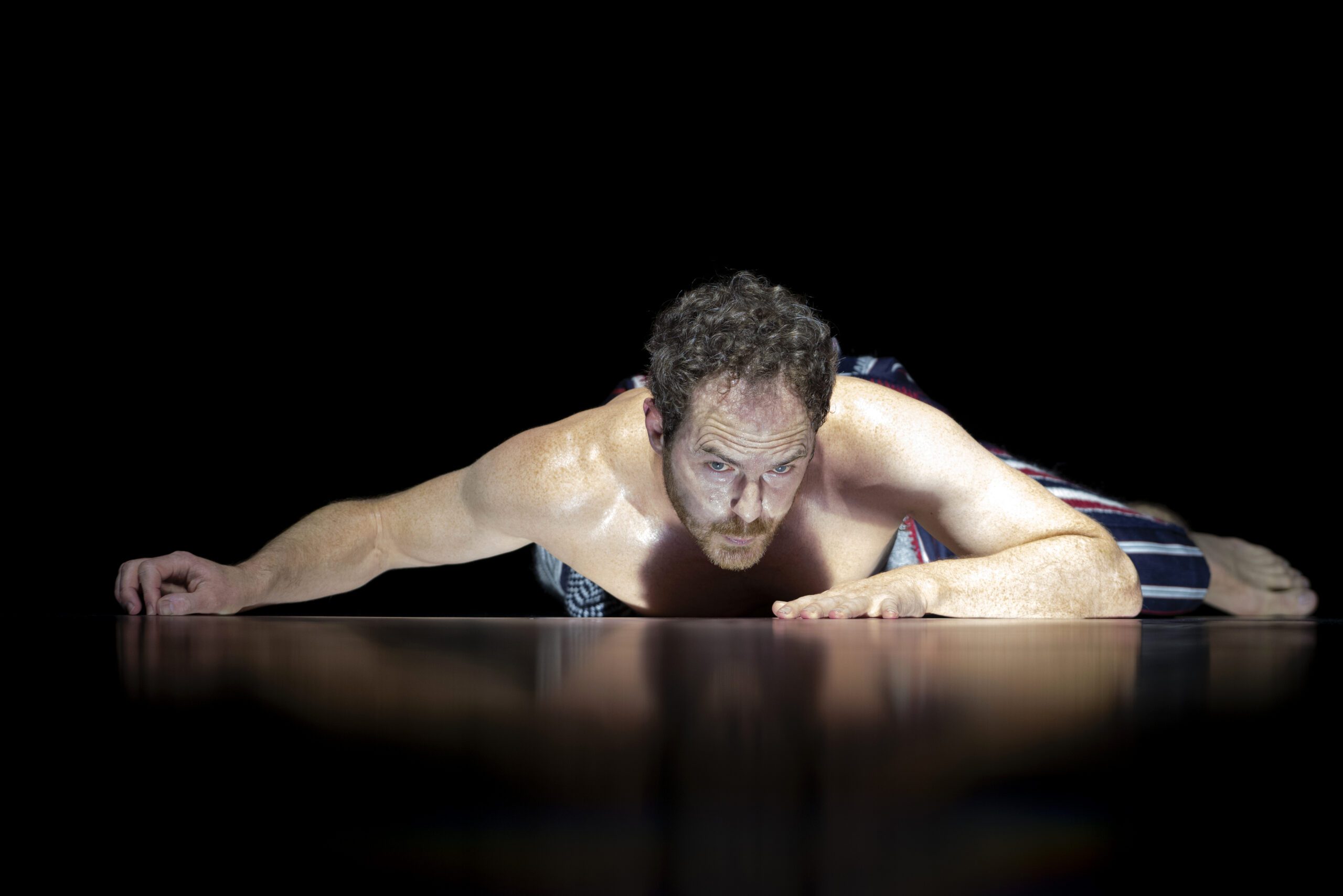 A man with curly hair and a beard lies on the floor, lit dramatically against a black background, with one arm extended forward. He appears intense and focused, with sweat visible on his skin.