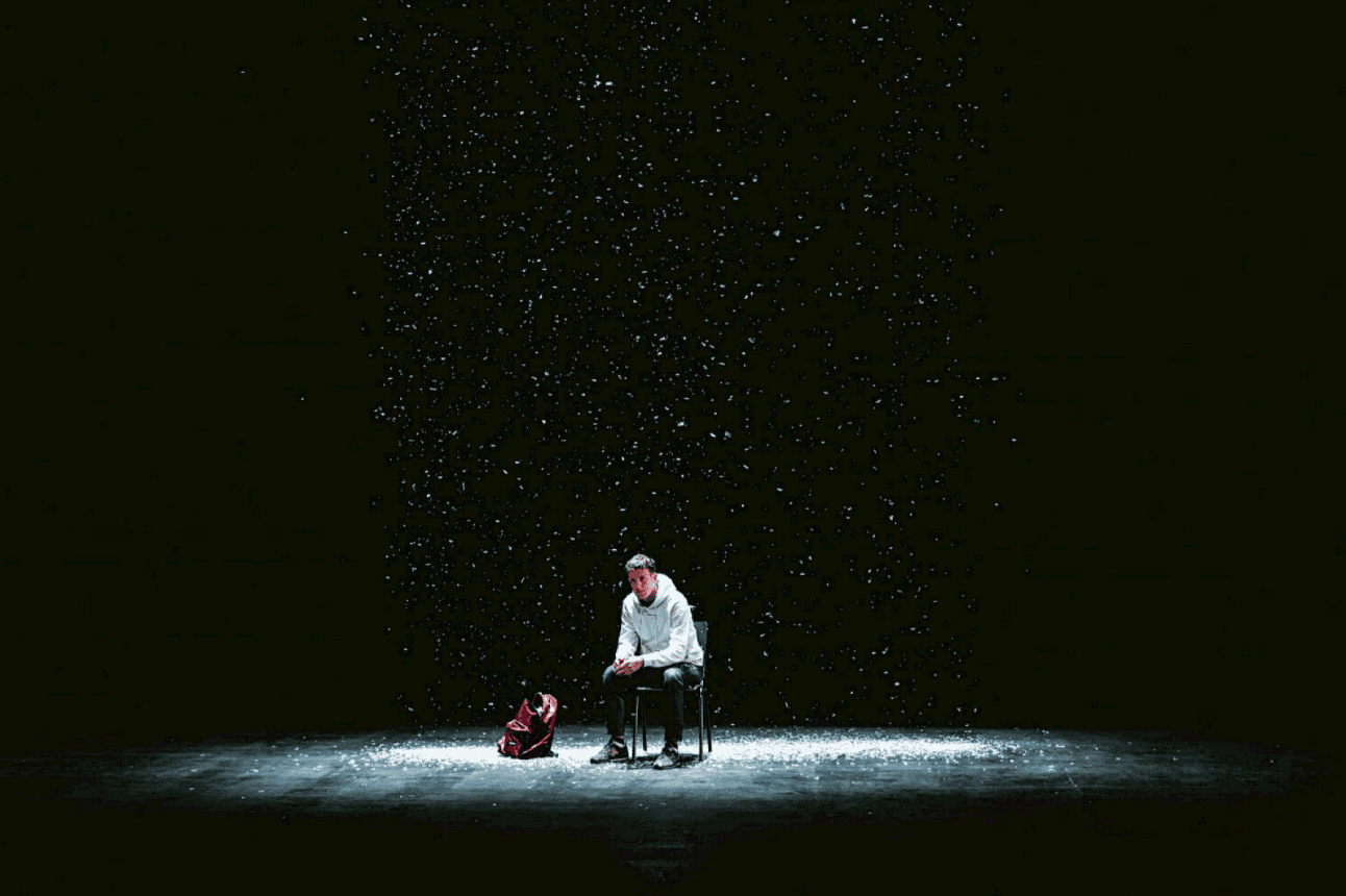 A person sits alone on a chair under falling snow, illuminated by a spotlight, with a red backpack on the floor beside them in a dark, empty space.