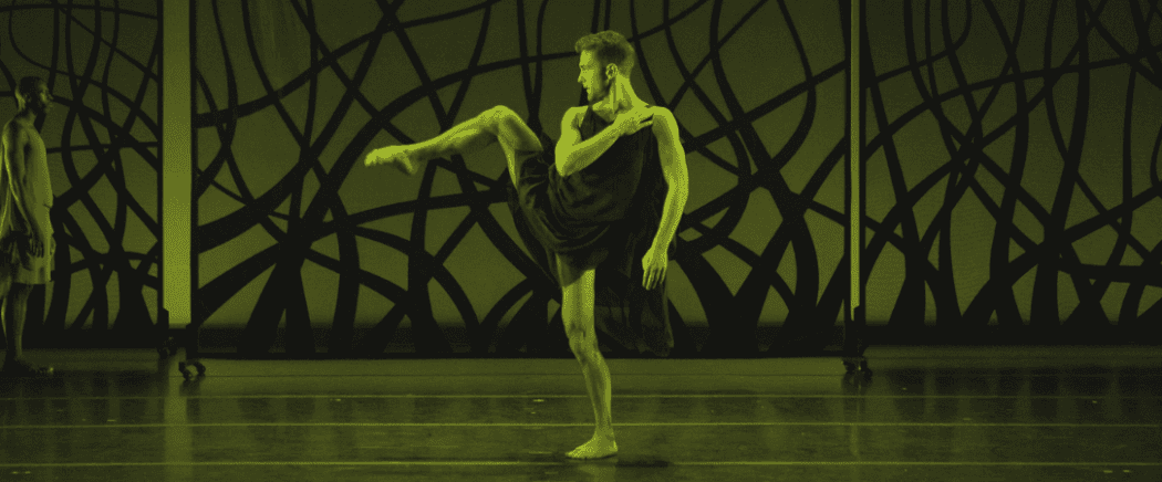 A dancer in a dark, flowing outfit performs on stage, balancing on one leg with the other leg raised, against an abstract, wavy-lined backdrop. Another person stands in the background. The image has a greenish tint.