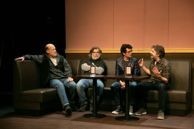 Four men sit in two booth seats around a wooden table, engaged in conversation. Three face forward, while one gestures animatedly. The setting appears to be a cafe or restaurant with dim lighting.