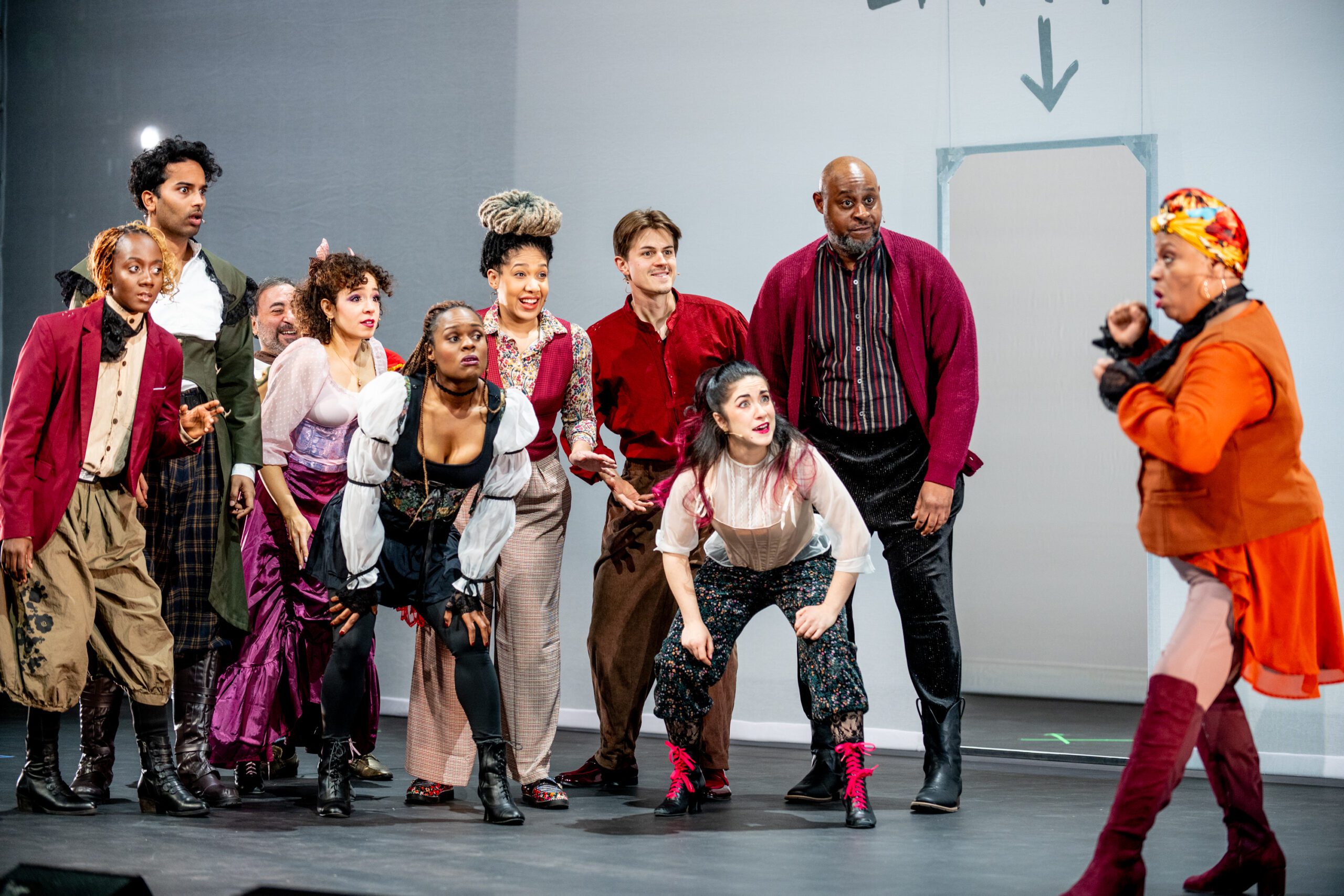 A group of actors in colorful costumes stand and squat on stage, watching a performer in orange speak animatedly; a gray wall and arrow are in the background.