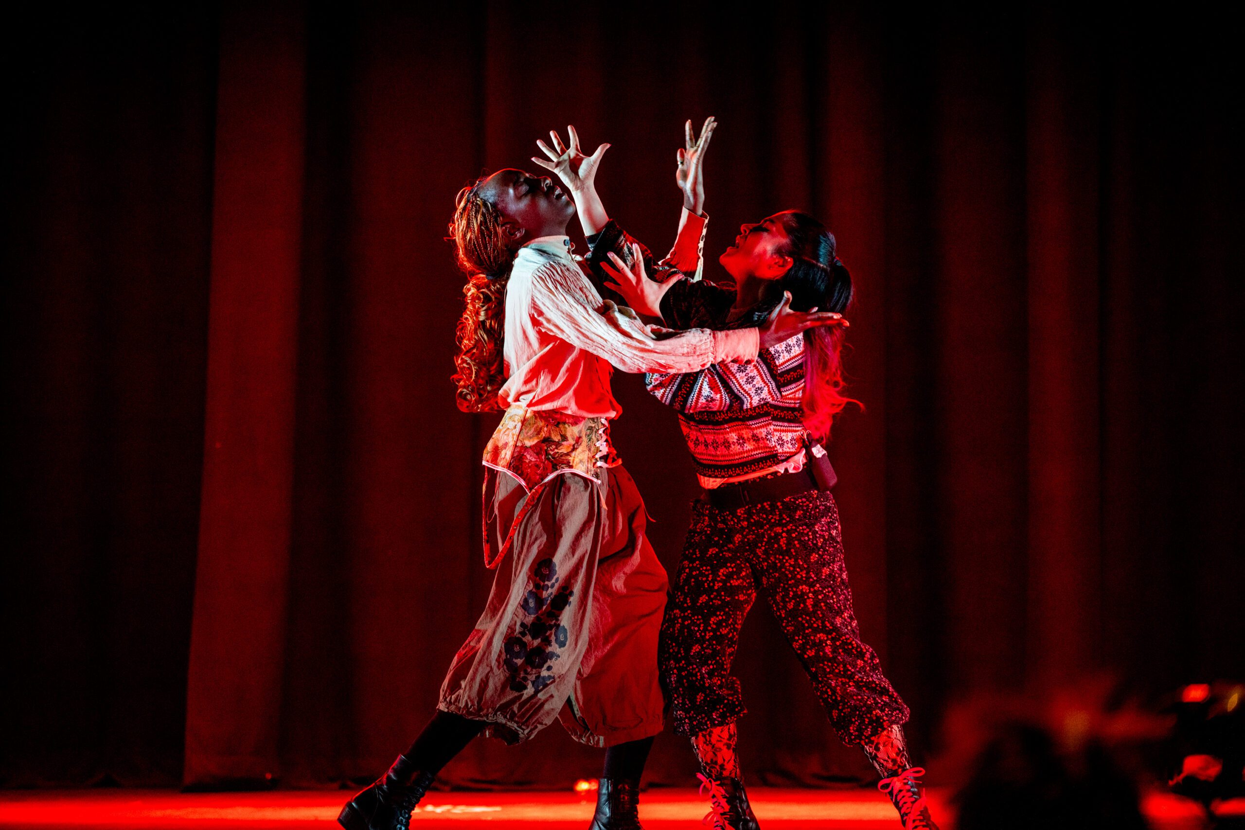 Two performers in patterned clothing pose dramatically on stage with red lighting and dark curtains, reaching their hands upward and toward each other, creating a sense of intensity and emotion.