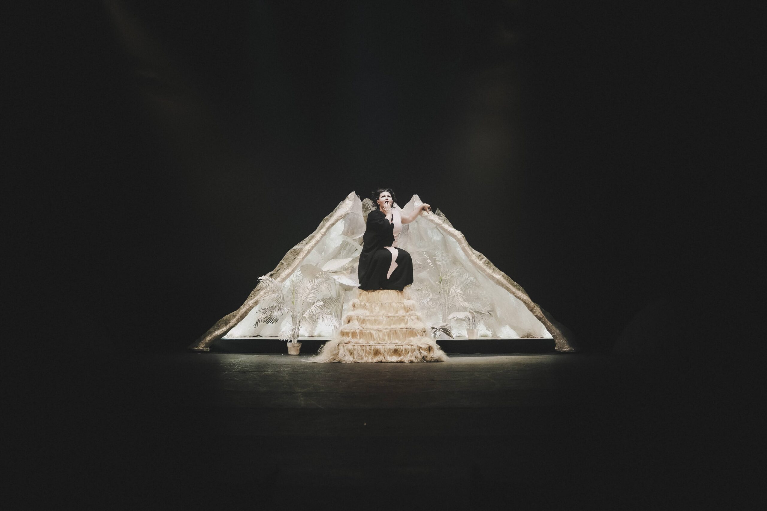 A performer in a dramatic costume sits center stage before a backdrop resembling a mountain, illuminated by spotlights against a dark background.