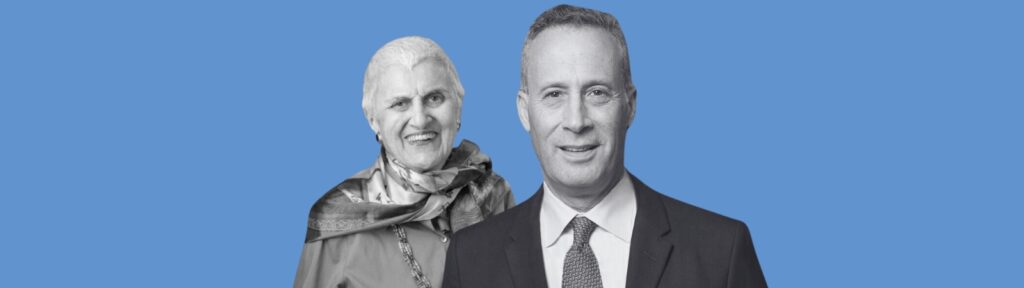An older woman wearing a patterned scarf and a man in a suit and tie stand in front of a solid blue background, both facing the camera and smiling.