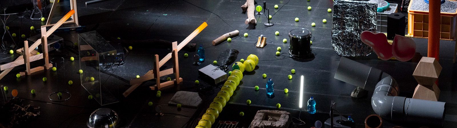 A variety of objects, including tennis balls, wooden planks, pipes, bottles, and foil, are scattered across a dark floor in an arrangement resembling a kinetic art installation.