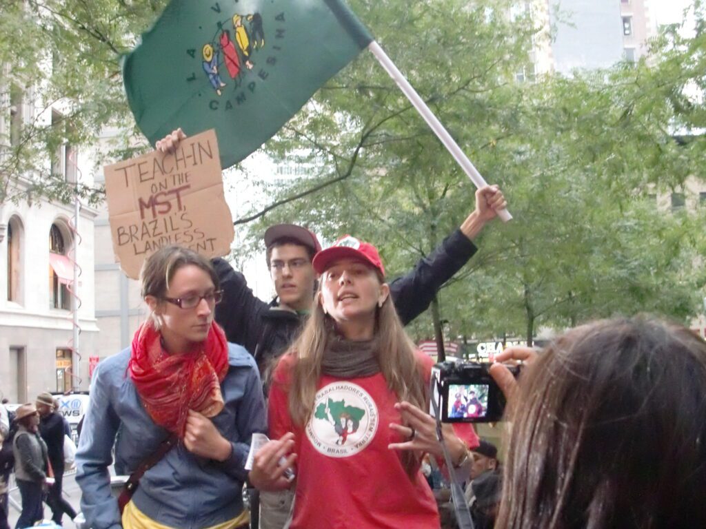 Three people stand outside; one holds a green flag, another holds a cardboard sign reading “Teach-in on the MST—Brazil’s Landless,” and a third person wears a red cap and shirt with a tree logo. A camera is visible in the foreground.