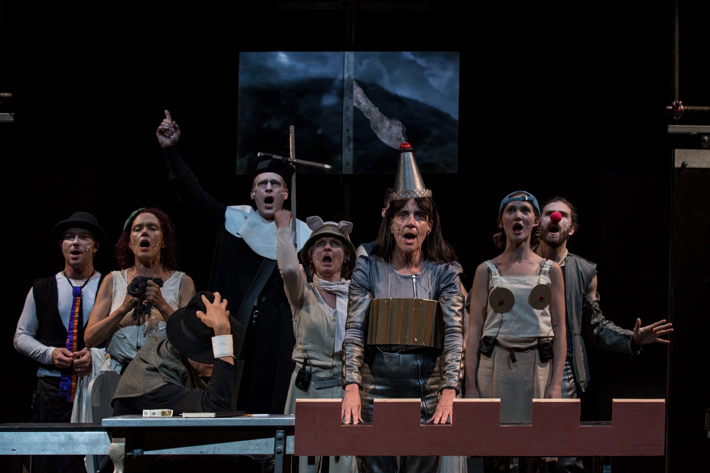 A group of actors in eclectic costumes perform dramatically on stage; some shout or gesture, while others wear hats or props, including a red clown nose and a cone hat, against a dark theatrical backdrop.