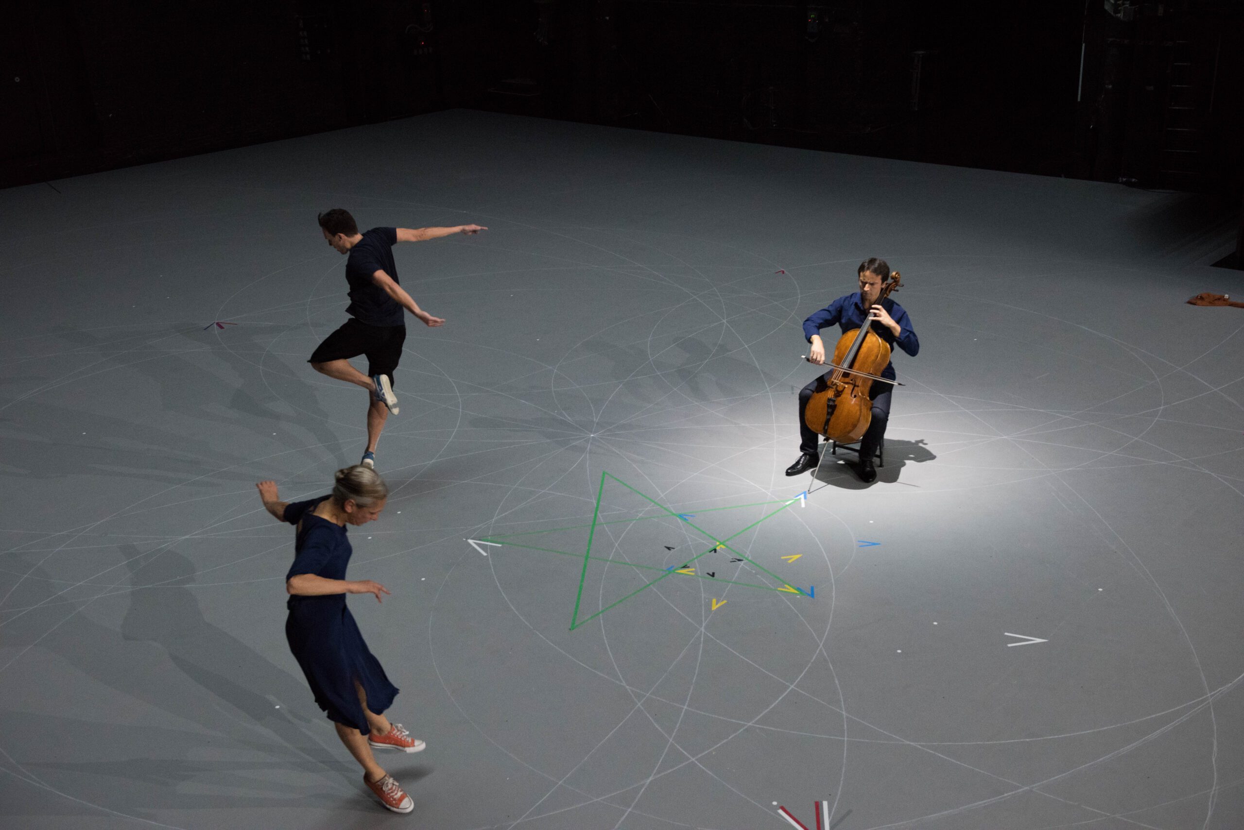 Three people perform on a gray stage with geometric patterns; one plays a cello under a spotlight, while two others dance nearby, surrounded by colored tape and scattered paper planes.