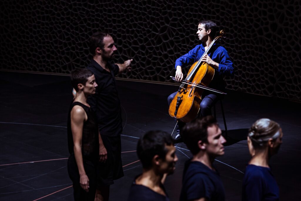 A cellist in a blue shirt plays on stage while several people stand in a line in the foreground; one person gestures, pointing toward the cellist. The background features a dark, textured wall.