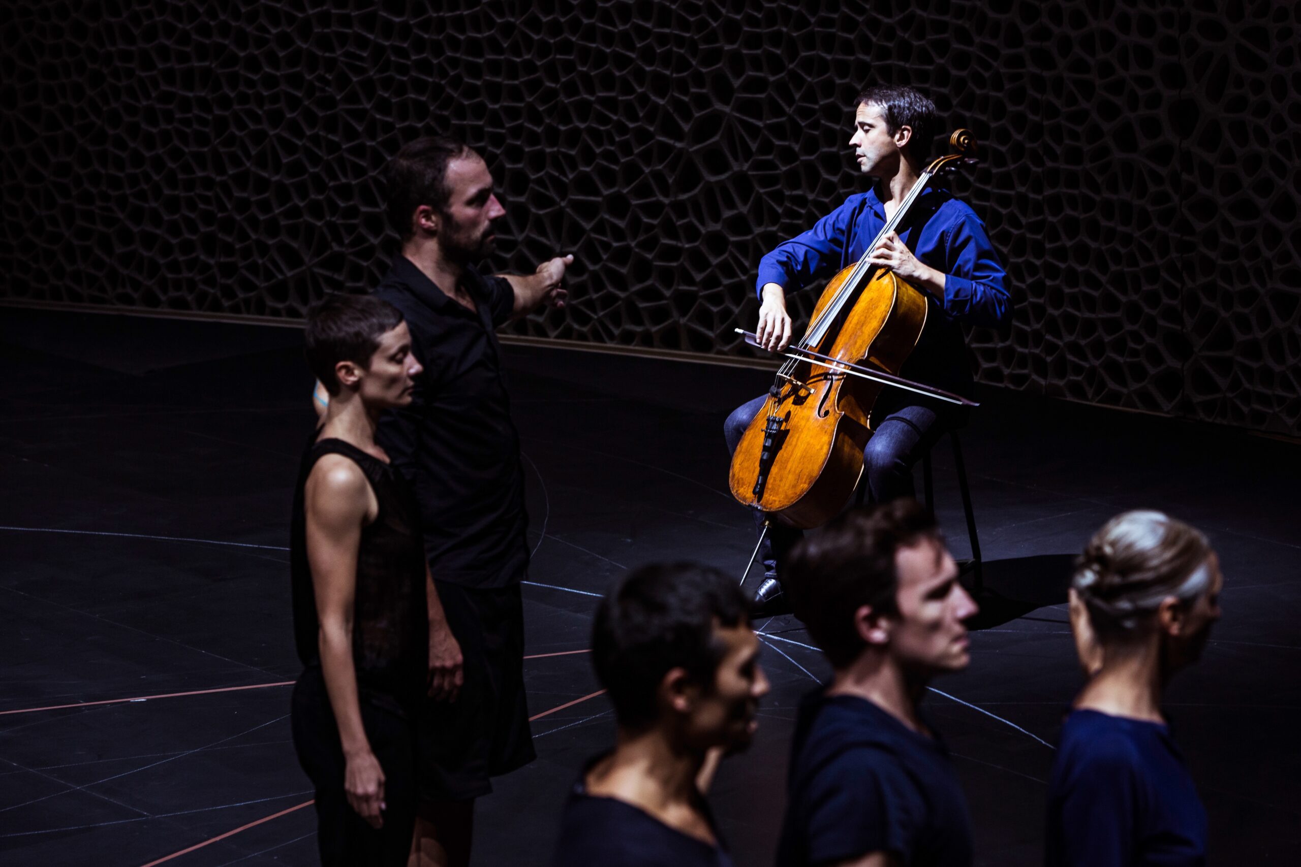 A cellist in a blue shirt plays on stage while several people stand in a line in the foreground; one person gestures, pointing toward the cellist. The background features a dark, textured wall.
