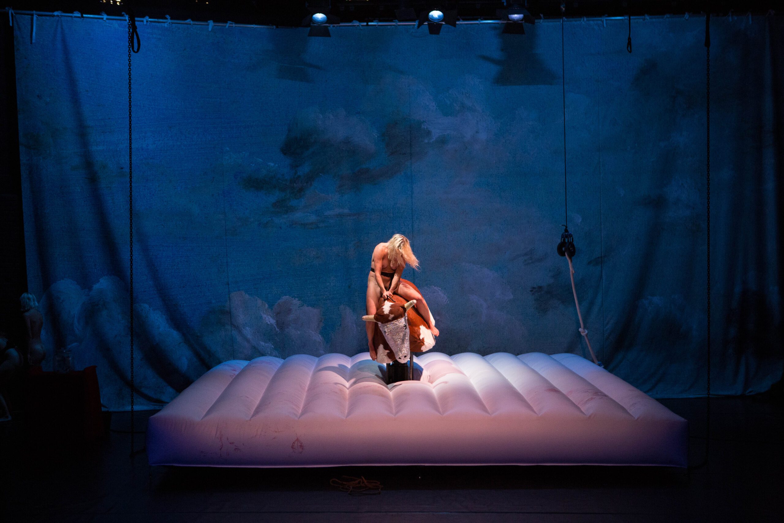 A person in light clothing stands on an inflated white mattress onstage, with blue cloud-patterned curtains behind them, under theatrical lighting.