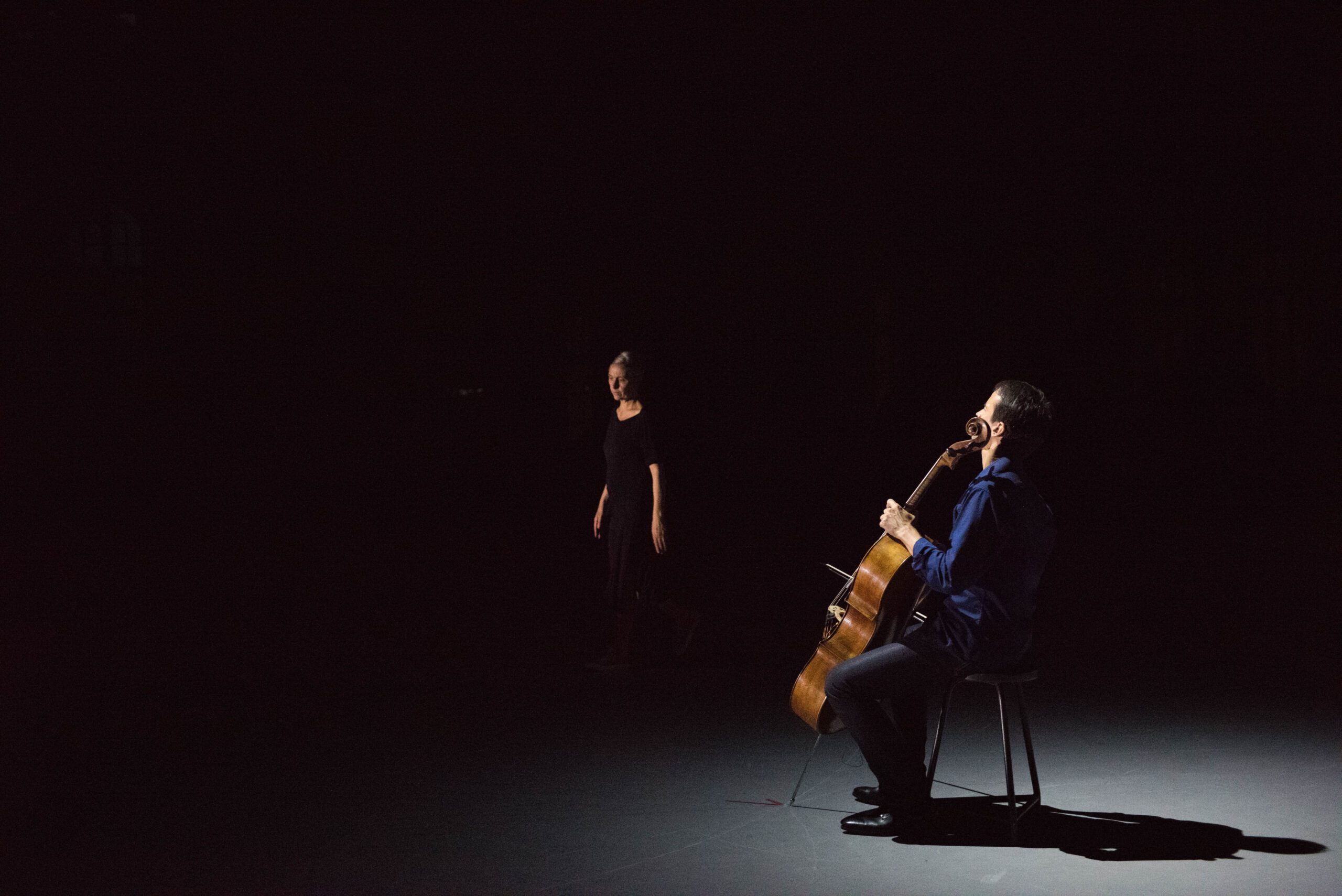 A cellist sits on a stool playing under a spotlight, while a person in dark clothing stands in the shadowy background on stage.
