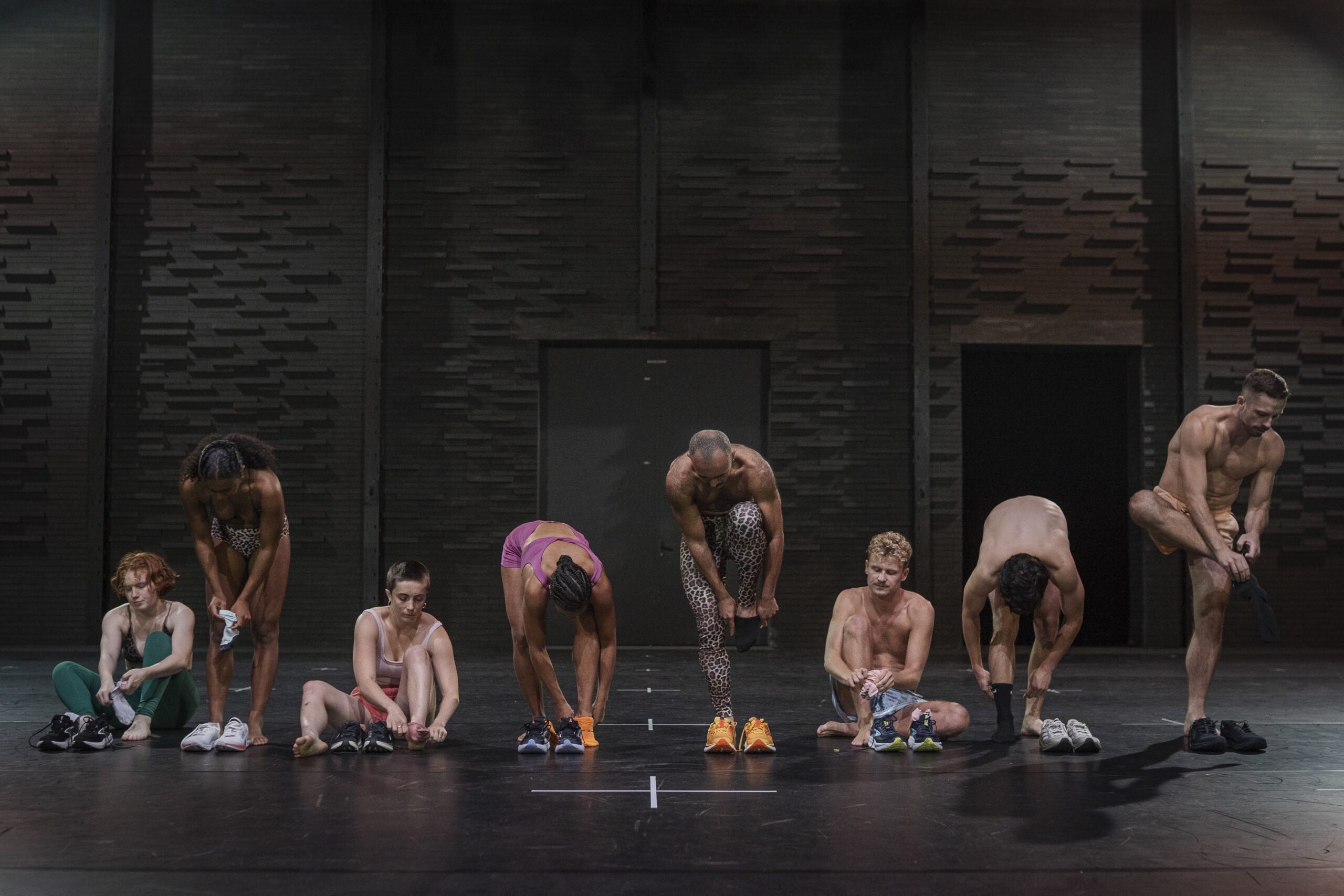 Seven people on a stage, all wearing patterned leggings, bend over as they change their shoes together, some standing and some sitting, against a dark, textured wall background.