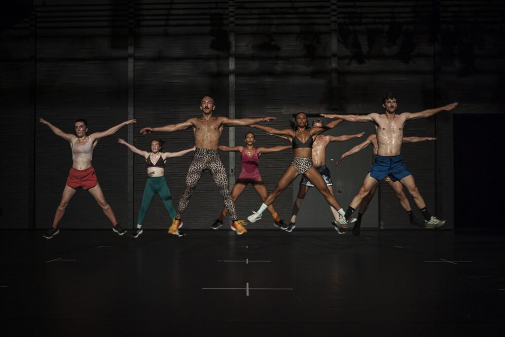 Seven people are mid-jump on a dimly lit stage, arms and legs spread wide in a star shape, all in athletic attire. The background is dark and industrial, with subtle lighting overhead.