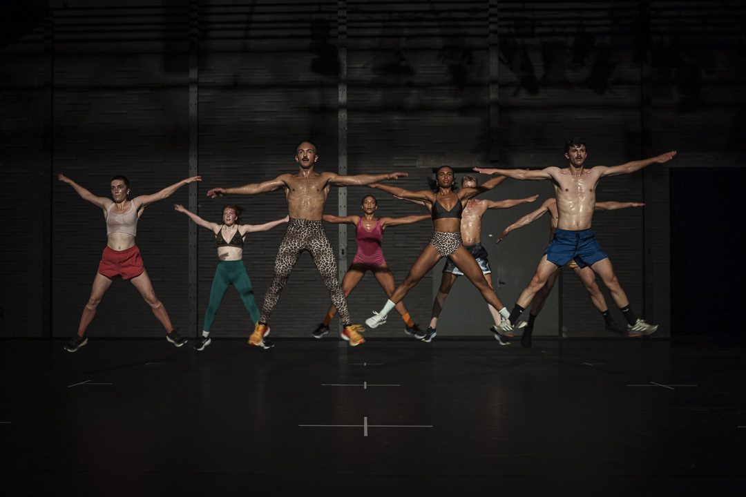 Seven people are mid-jump on a dimly lit stage, arms and legs spread wide in a star shape, all in athletic attire. The background is dark and industrial, with subtle lighting overhead.