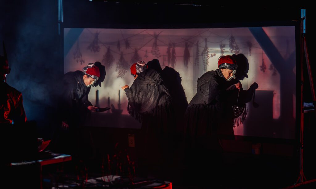 Three people in dark, dramatic costumes and red headscarves perform on stage with eerie lighting. Shadows and dried herbs hang in the background, creating a mysterious atmosphere.