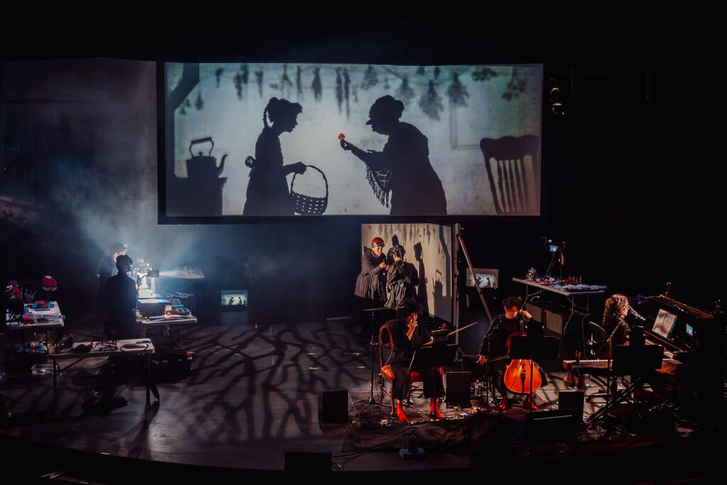 A shadow puppet show is projected above a stage where musicians play string instruments, surrounded by props and equipment, with dramatic lighting and silhouettes cast on the floor.