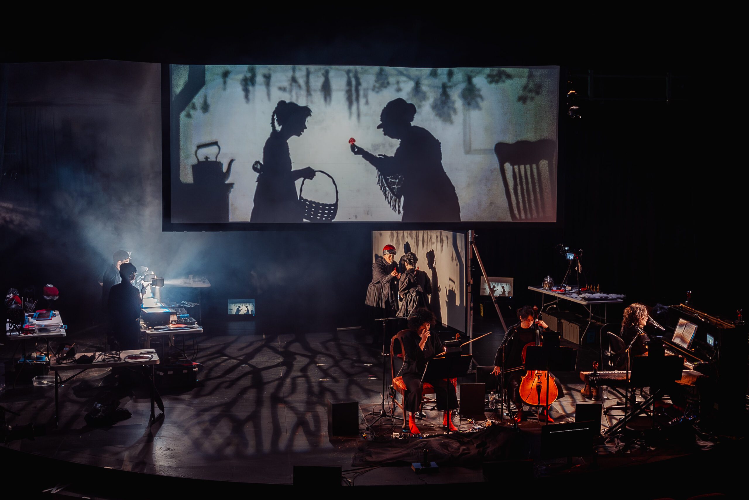 A shadow puppet show is projected above a stage where musicians play string instruments, surrounded by props and equipment, with dramatic lighting and silhouettes cast on the floor.