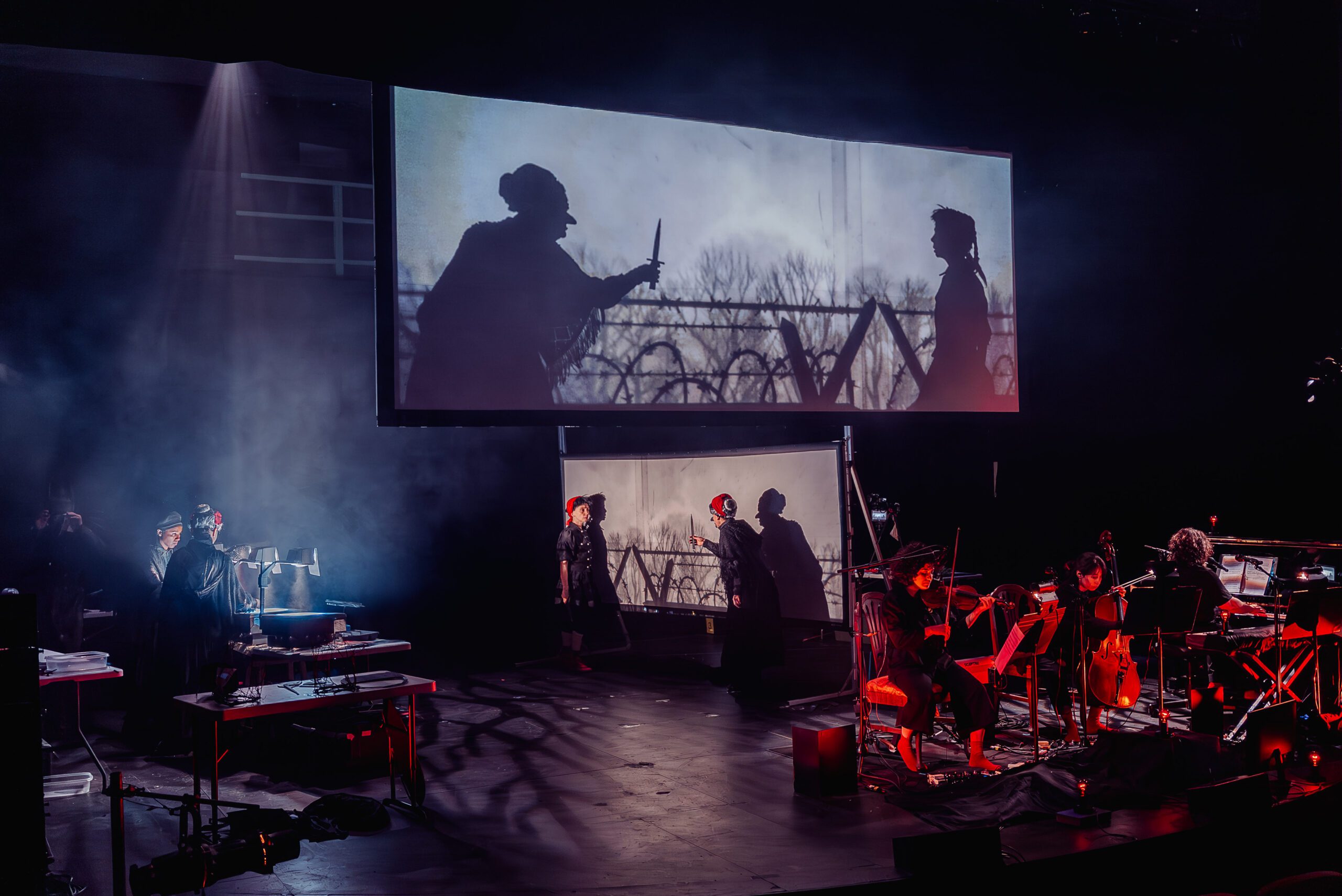 A stage performance with musicians playing string instruments, actors in costume, and a large screen displaying dramatic silhouettes, all under moody, colorful lighting.