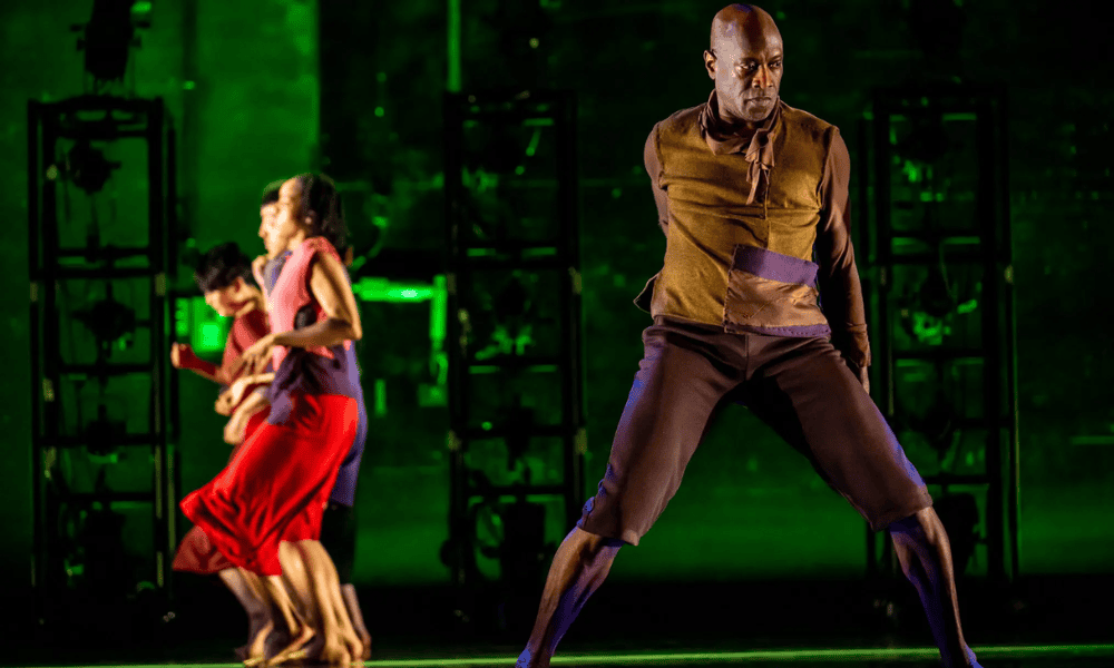 Four dancers perform on stage with dramatic lighting; the foreground features a man in brown and purple attire striking a powerful pose, while three dancers in colorful outfits move energetically behind him.