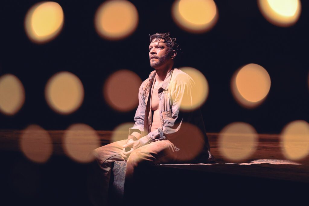 A man sits alone on a bench in dim lighting, with soft, out-of-focus circular lights in the foreground creating a dramatic, moody atmosphere.