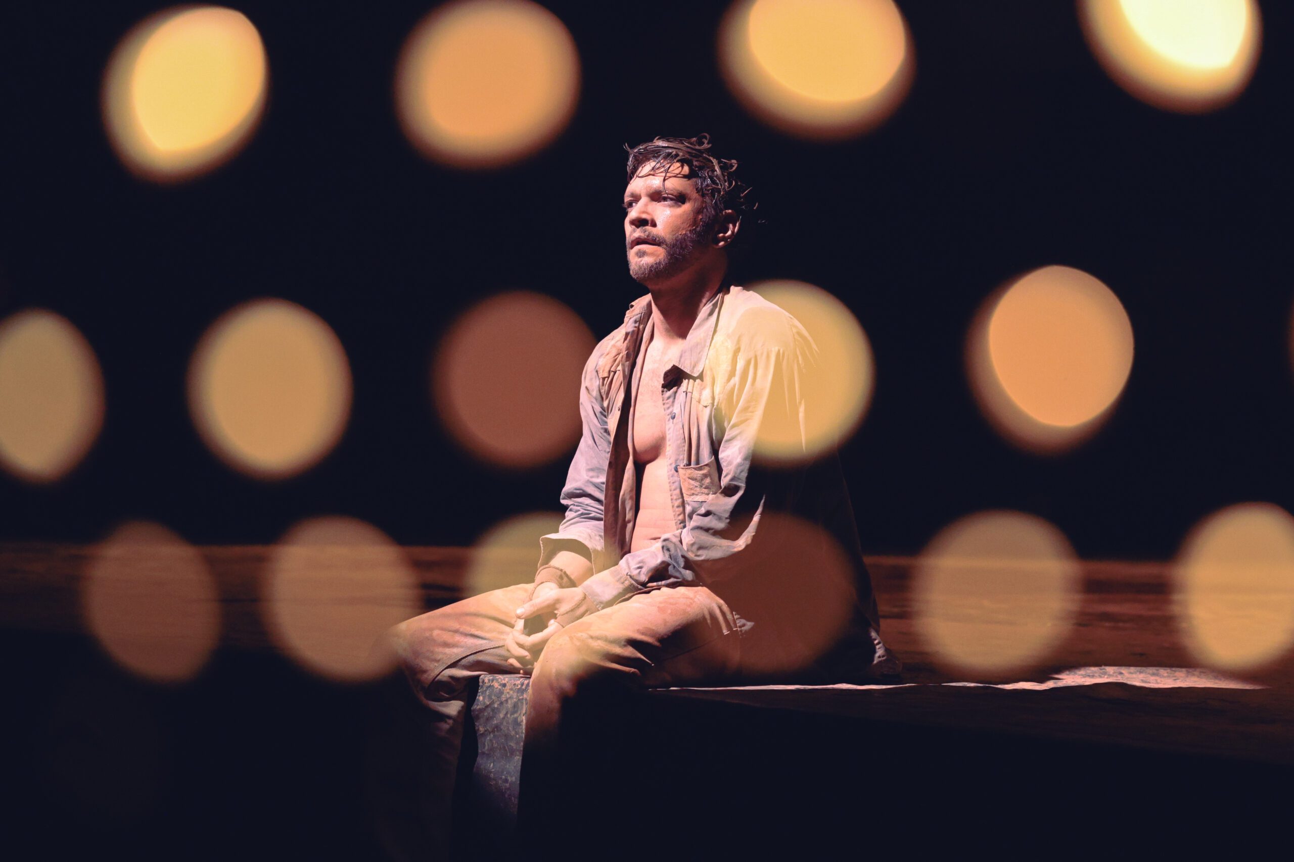 A man sits alone on a bench in dim lighting, with soft, out-of-focus circular lights in the foreground creating a dramatic, moody atmosphere.
