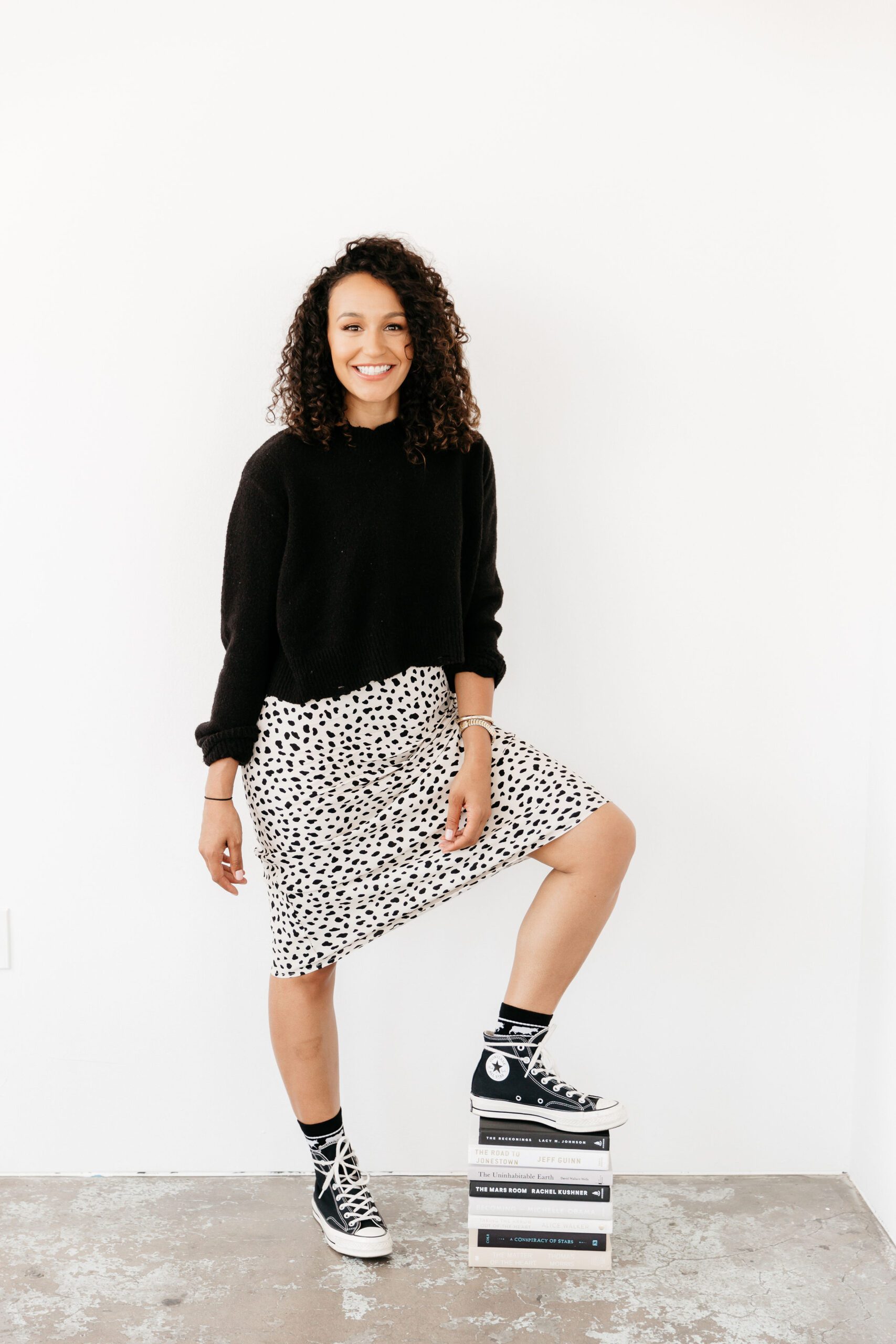 A smiling woman with curly hair wearing a black sweater and spotted skirt, standing with one foot resting on a stack of books.