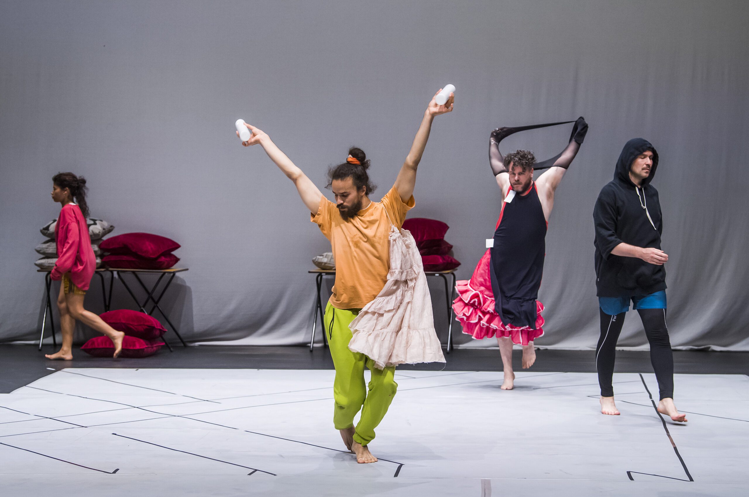Four people perform on stage; one in orange shirt and green pants poses with arms up, another in a dress holds fabric, a third in black hoodie walks, and a fourth stands near stacked pillows. The background is plain and gray.