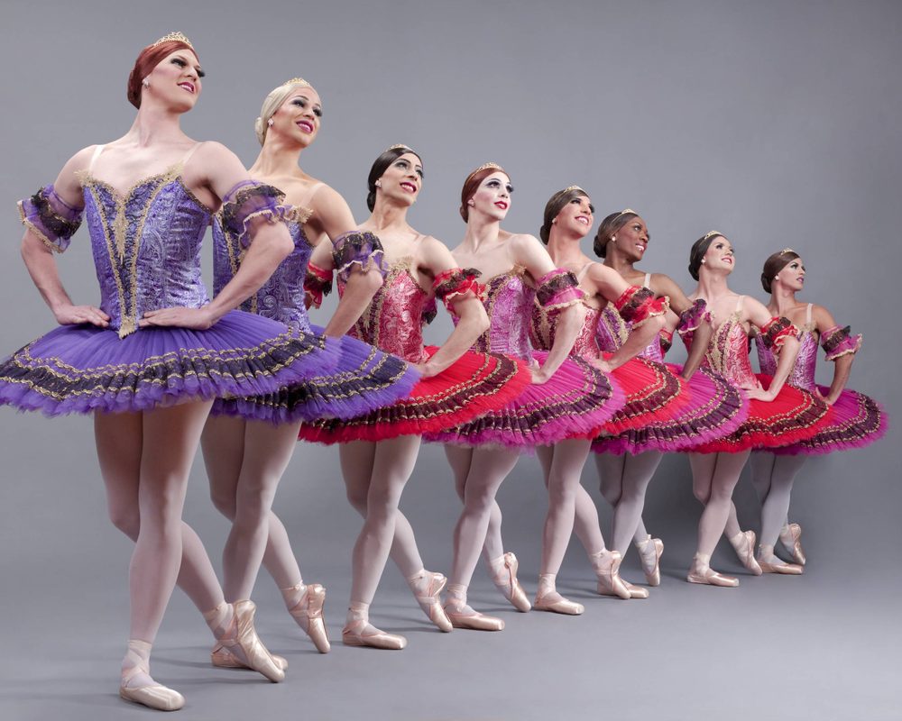 Seven ballet dancers in colorful tutus, standing en pointe in a line, hold hands and smile. Their tutus range from purple to various shades of pink and red, and they pose against a neutral gray background.