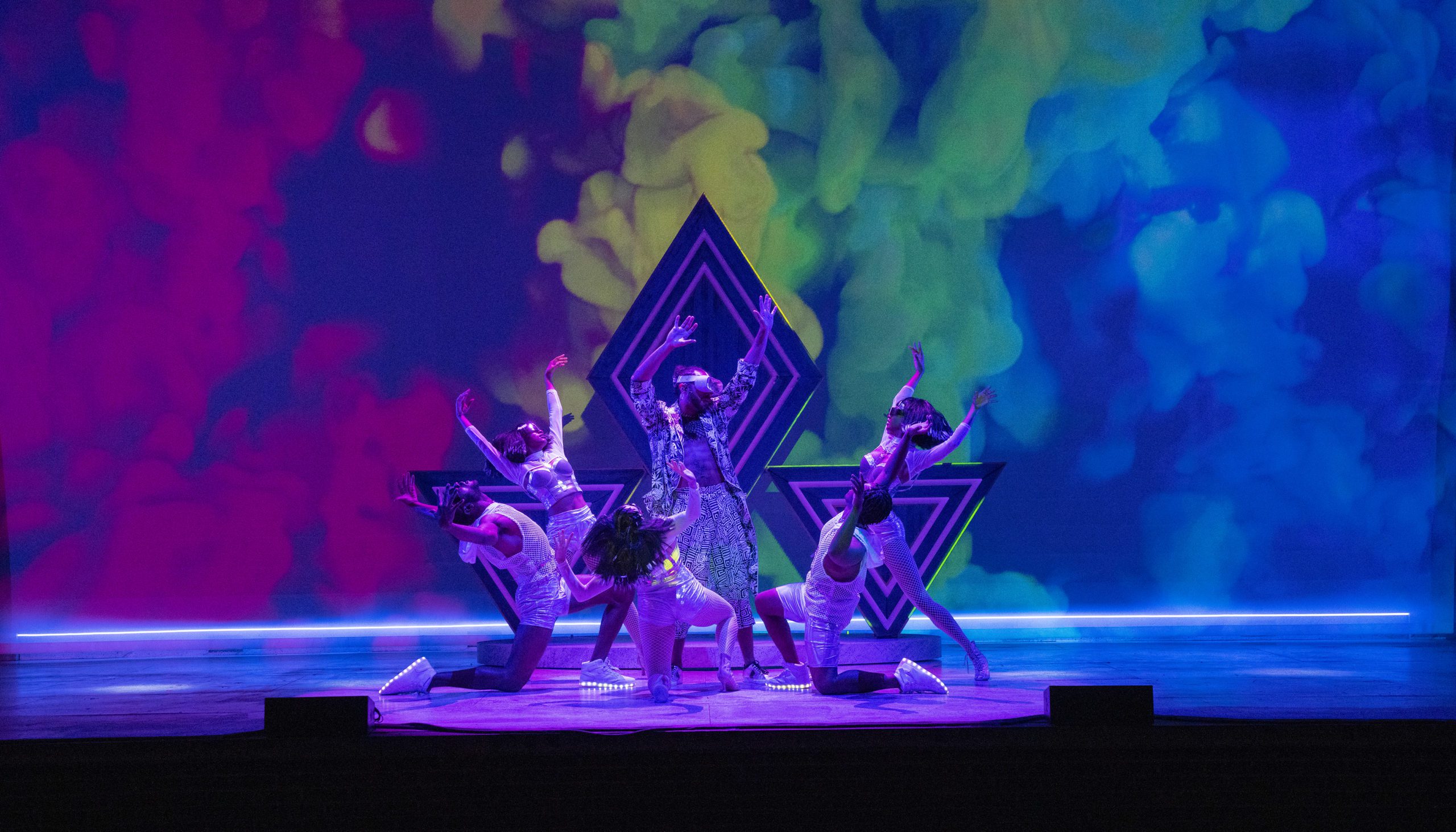 Six performers pose dramatically on stage with vibrant, swirling colors projected in the background. Geometric shapes and bright lighting enhance the theatrical, energetic atmosphere.
