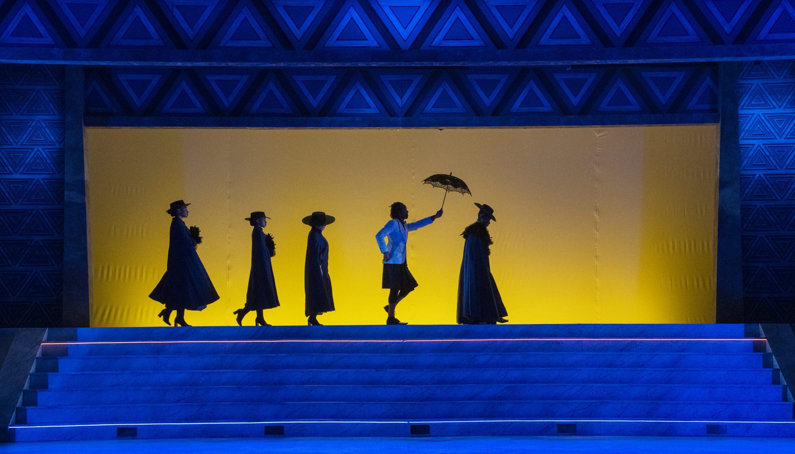 Five people in silhouette stand on stage; four wear hats and coats, while one in the center holds an umbrella aloft. The background glows yellow, framed by a blue, geometric arch.