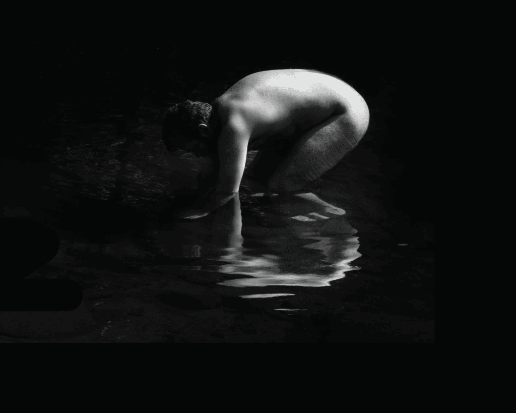 A nude person crouches at the edge of water, their reflection visible on the surface. The image is in black and white, with dramatic lighting highlighting the figure against a dark background.