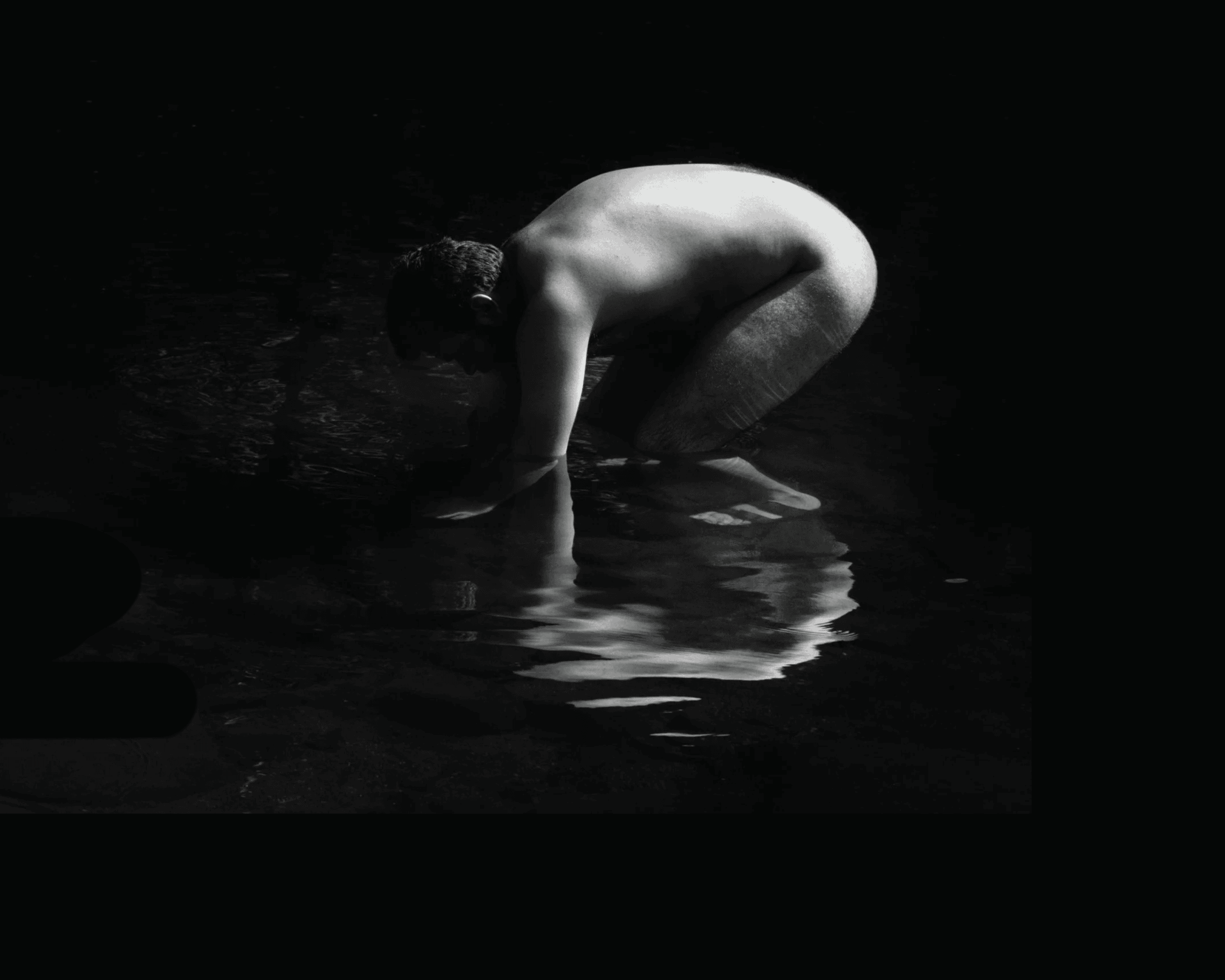 A nude person crouches at the edge of water, their reflection visible on the surface. The image is in black and white, with dramatic lighting highlighting the figure against a dark background.
