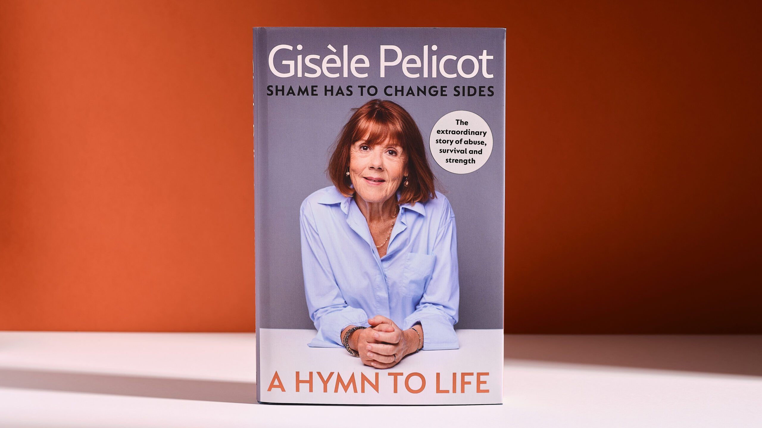 The book “A Hymn to Life” by Gisèle Pelicot features a portrait of an older woman in a blue shirt sitting at a white table, with a warm orange background.