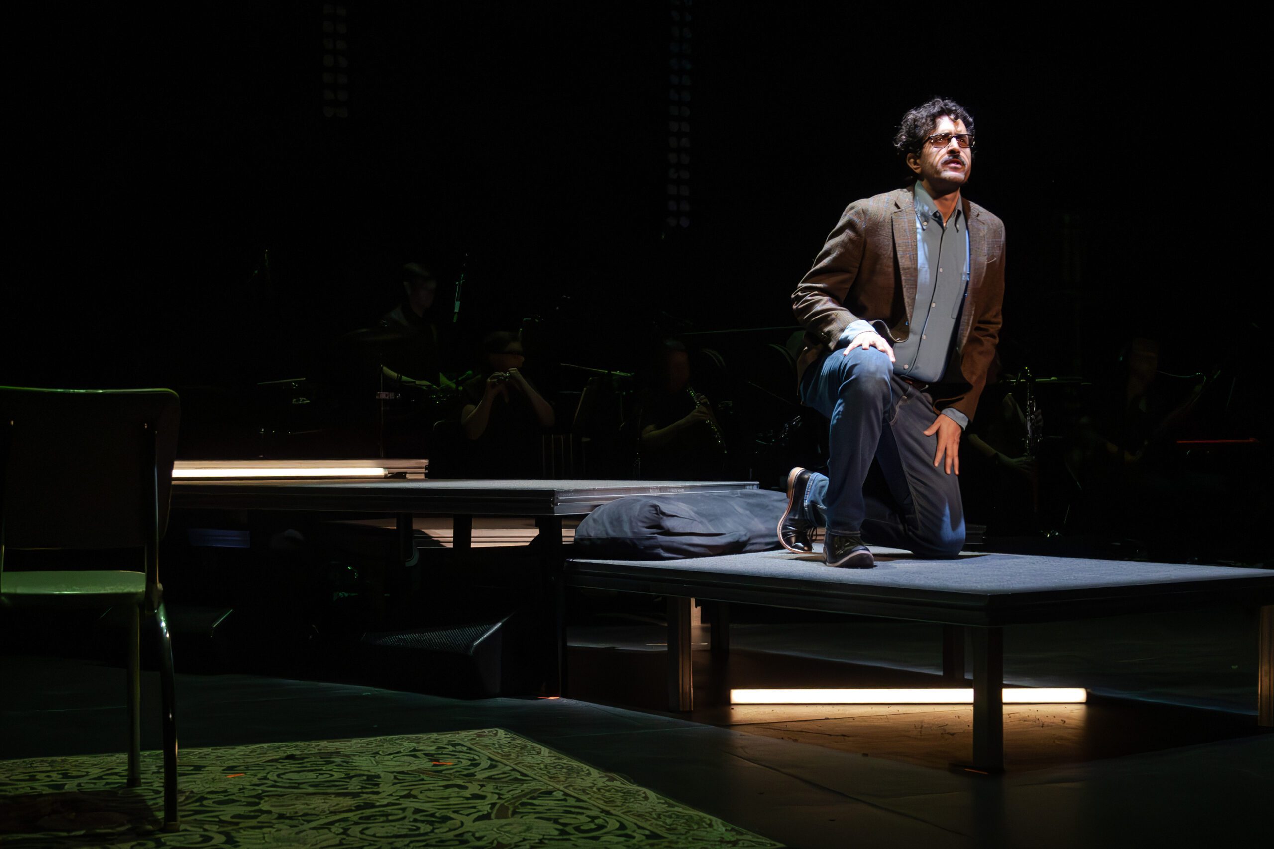A man in glasses, a blazer, and jeans kneels on a dimly lit stage platform, looking up. Musicians are faintly visible in the dark background. A chair and a patterned rug are also on stage.